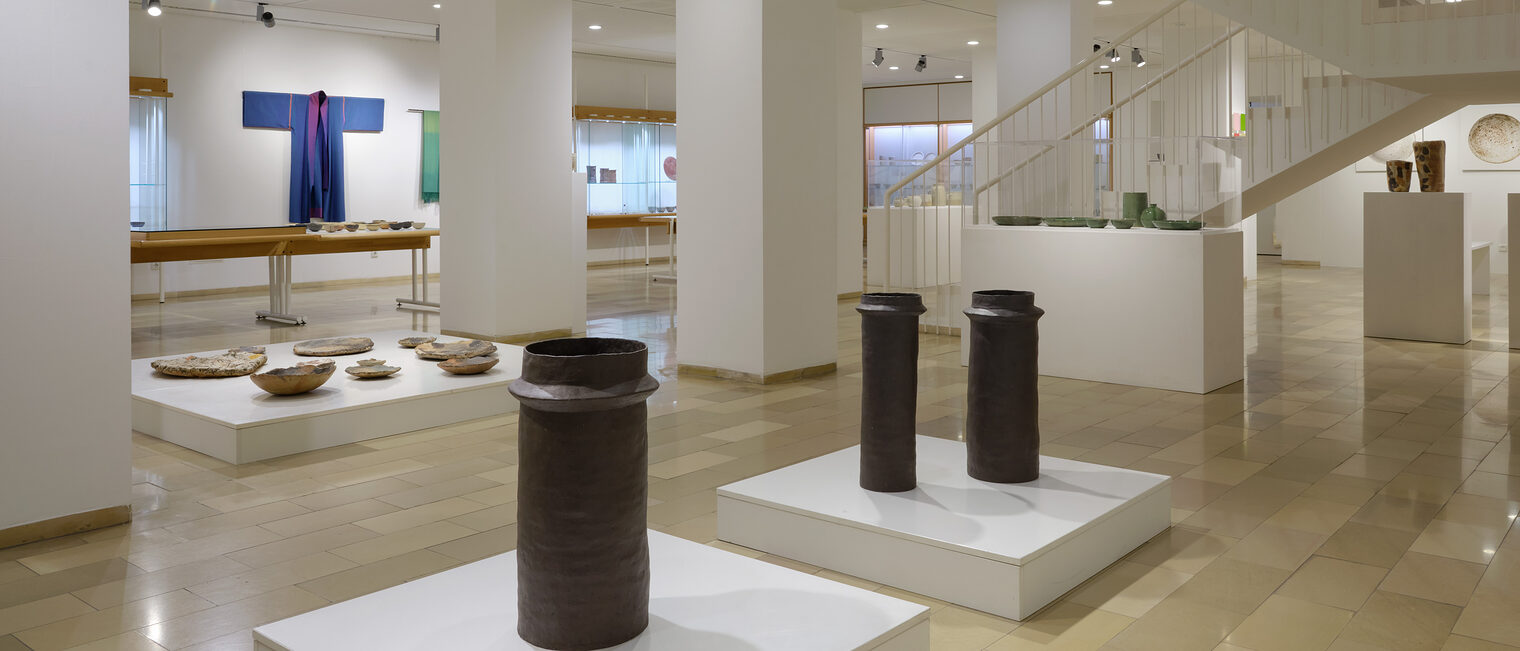 Untergeschoß der Galerie Handwerk mit zahlreichen Tonobjekten auf Podesten und kimono-artigen Objekten aus Seide