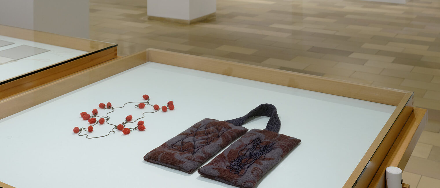 Zwei Objekte auf einem Tisch. Beide stellen in abstrakter Weise Schmuck aus Schnüren und Textilien dar.