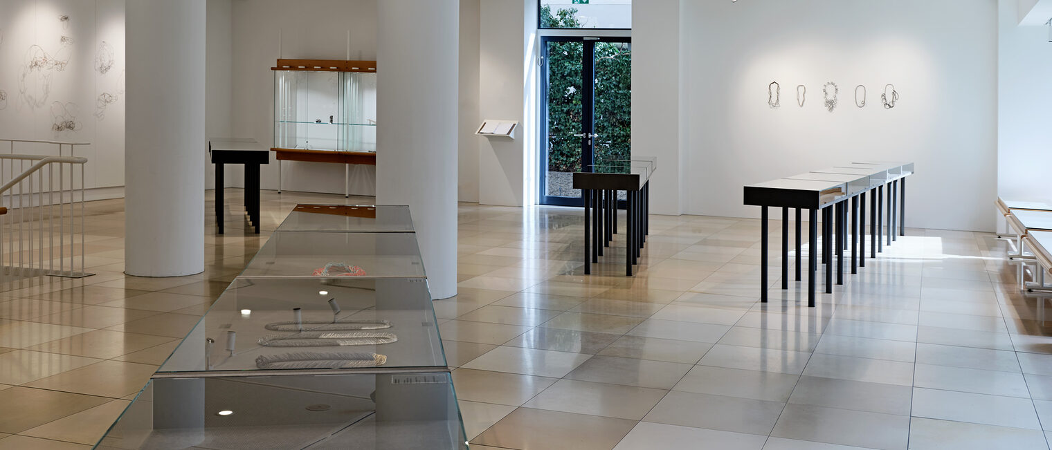 Einblick in die Galerie Handwerk im Kunstblock München während der Ausstellung Schmuck auf ganzer Linie mit Vitrinen und Tischen. 
