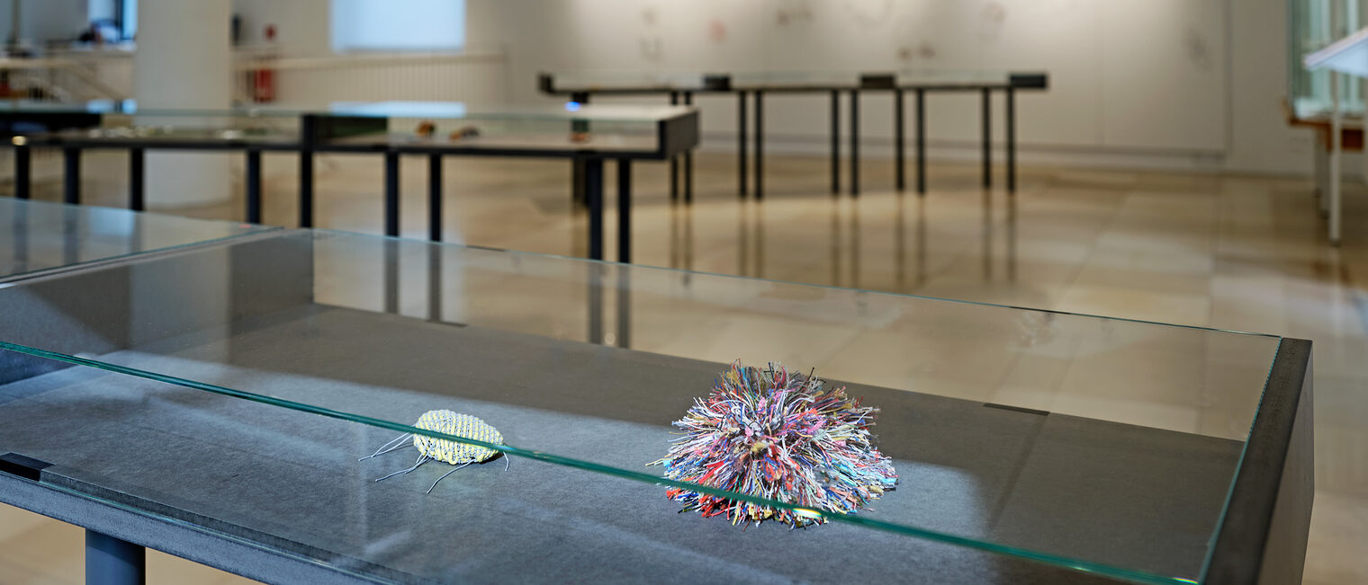 Blick in den Ausstellungsraum. Vitrine im Vordergrund zeigt ein buntes Gebilde aus Schnüren, die in alle Richtungen abstehen.