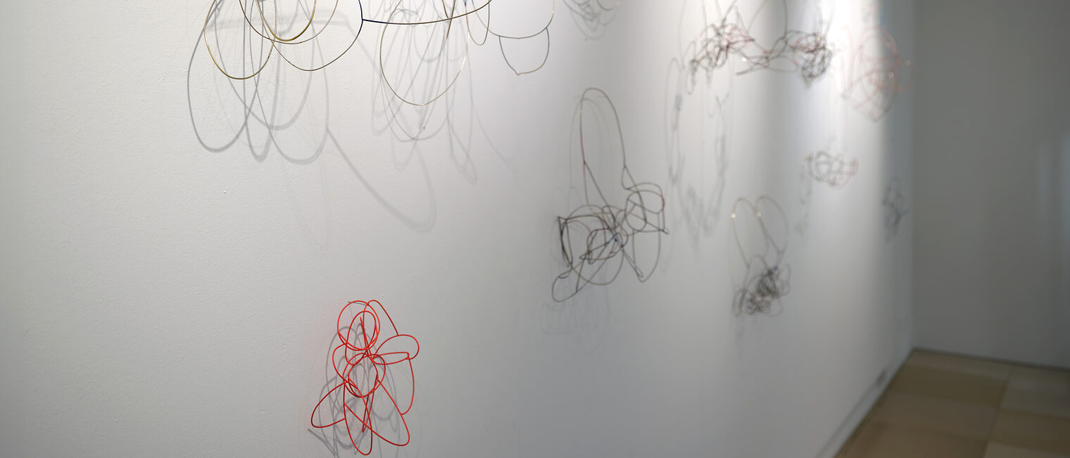 Mehrere abstrakt gebogene Objekte aus silbernem Draht an der Wand, eines davon in Signalrot, mit entsprechenden Schatten.