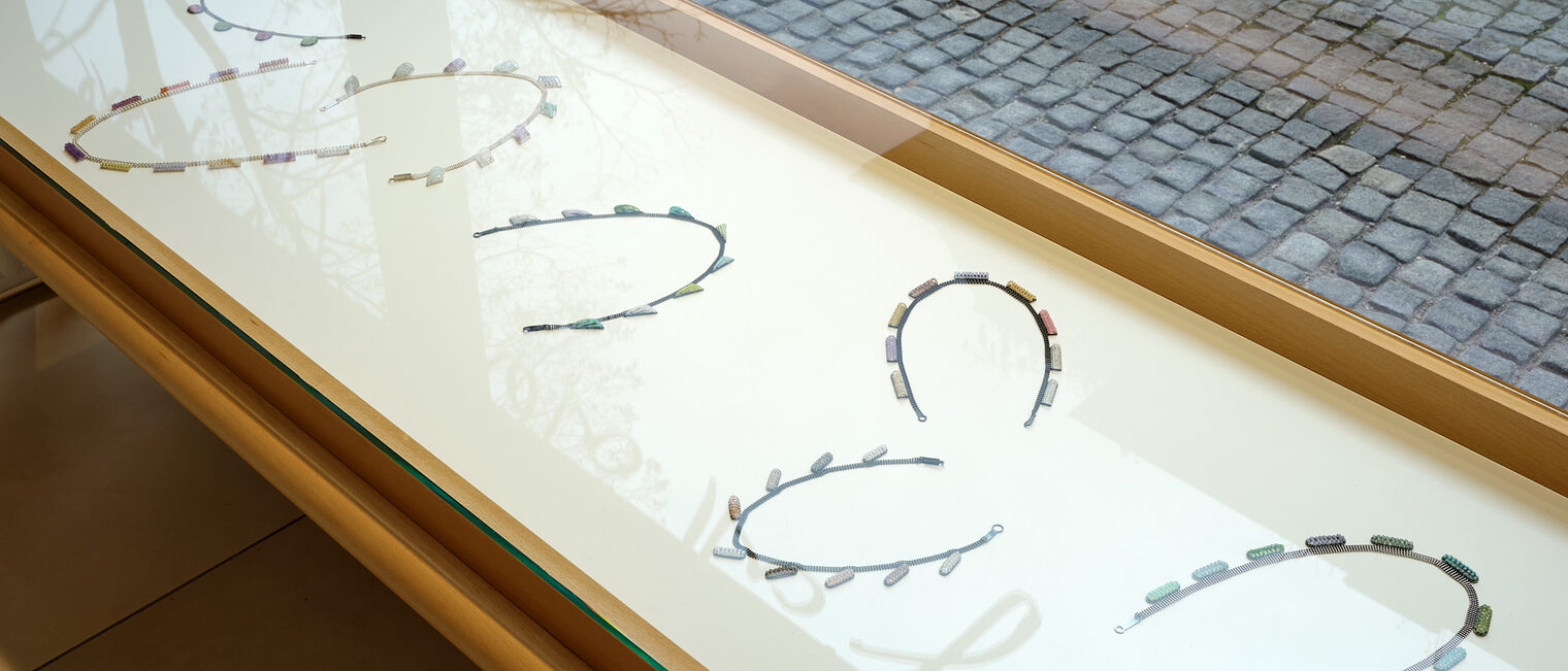 7 Ketten im gleichen Stil in einer Tischvitrine vor dem Schaufenster der Galerie Handwerk mit Blick auf Kopfsteinpflaster