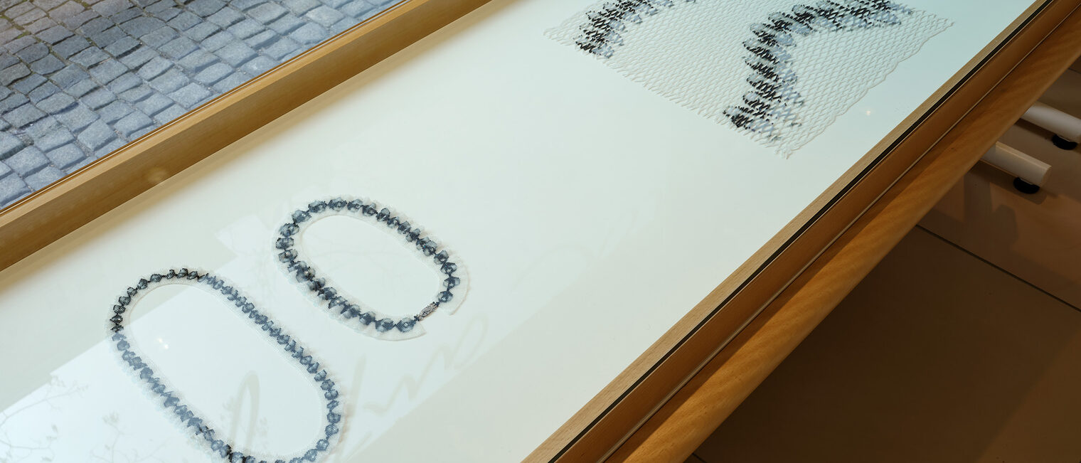 mehrere 2-dimensionale Objekte in grau, die Ketten darstellen in Tischvitrinen vor dem Schaufenster der Galerie Handwerk.