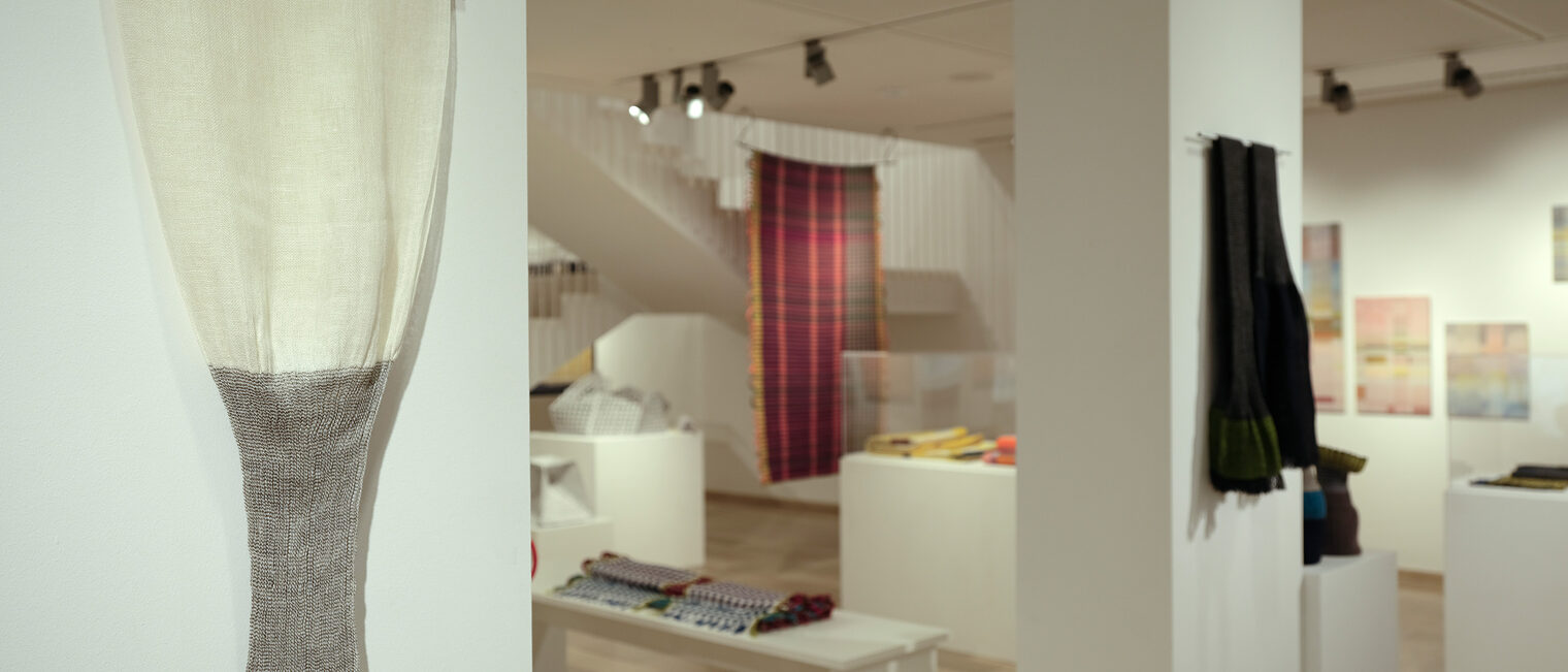 Blick durch den Galerieraum mit bunten, gewebten Plaids an Wänden und von der Decke hängend, in der Ausstellung Weben.