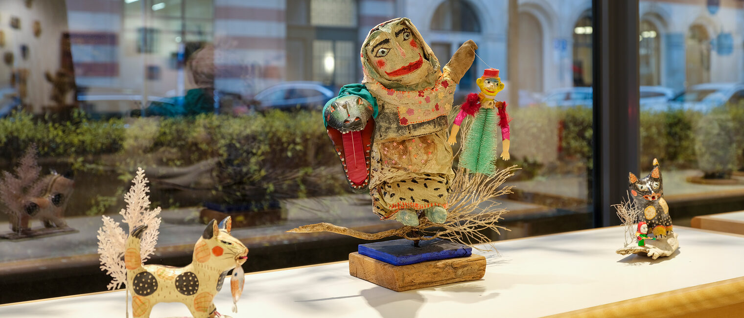 Auf der Ablage vor dem Schaufenster ist eine stimmungsvolle Szene mit Tieren und einer Kräuterhexe aus Holz dargestellt.